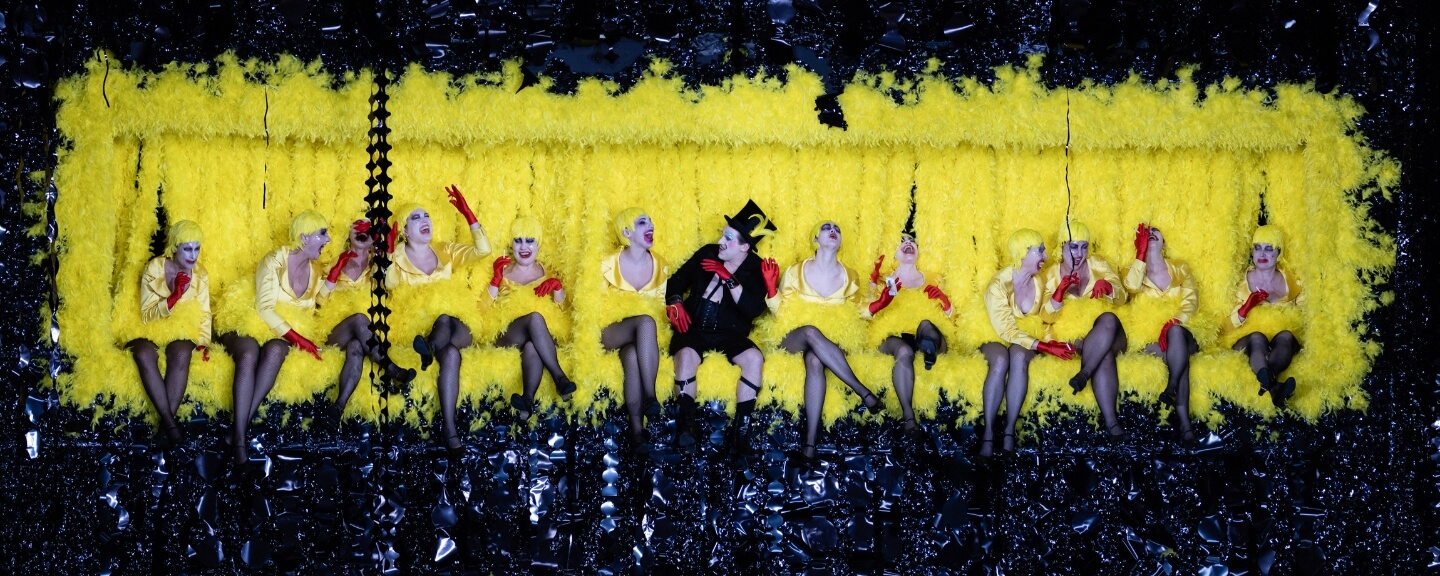 Row of women with a man in the middle against a yellow backdrop - Photo from The Cunning Little Vixen opera.